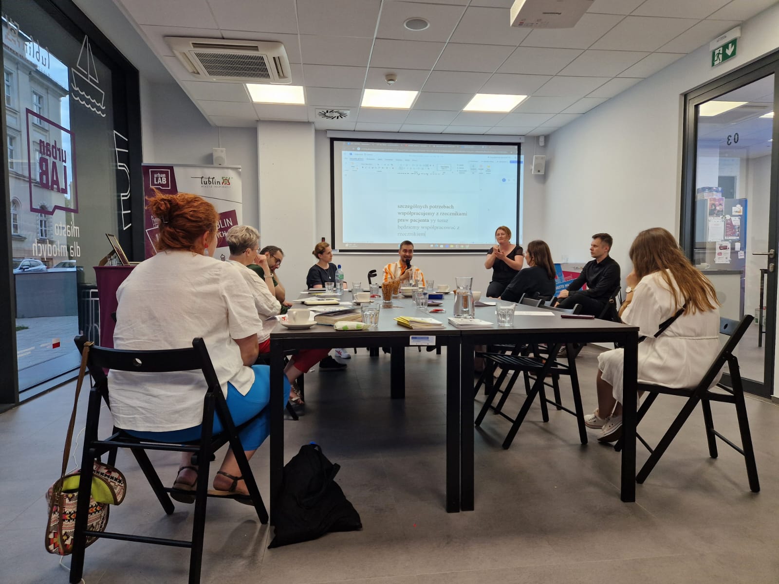 Grupa osób uczestniczy w warsztatach. Siedzą wokół stołu. Z przodu ekran rzutnikiem. Widać tłumaczy PJM.