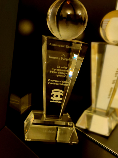 Szklana statuetka w formie odwróconego ostrosłupa z kulą z napisem "Ambasador dostępności. Pan Tomasz Wojakowski". poniżej uzasadnienie wyboru i logo Fundacji Widzialni.
