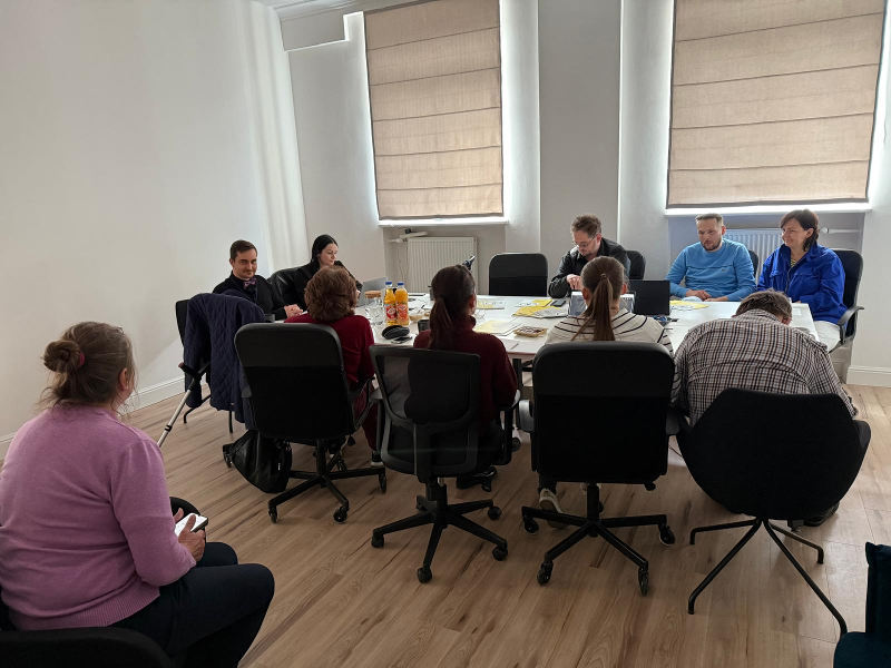 W jasnym pomieszczeniu biurowym grupa osób siedzi wokół owalnego stołu, prawdopodobnie biorąc udział w spotkaniu lub dyskusji. Większość osób jest zwrócona w stronę stołu.