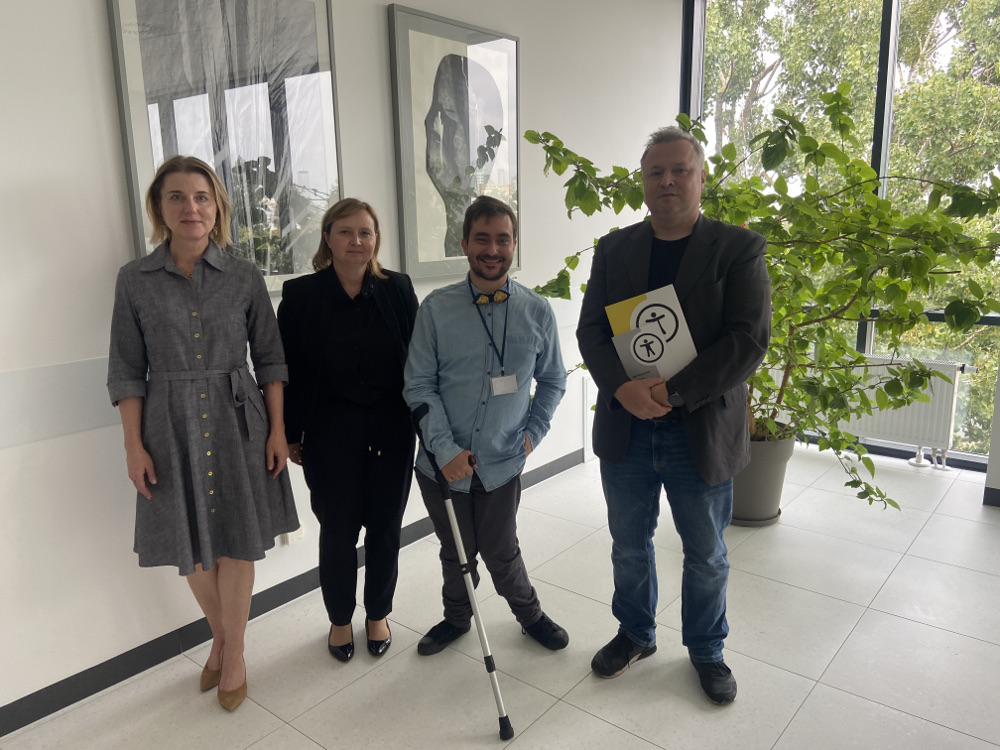 Cztery osoby stoją w jasnym korytarzu przy ścianie z obrazami i dużym oknem. Obok (lekko z tyłu) stoi zielona roślina w donicy. Osoby stoją obok siebie. Dwie kobiety i dwóch mężczyzn. Mężczyzna w jasnej koszuli z białą laską znajduje się pośrodku. Obok niego, po prawej, mężczyzna w ciemnej marynarce trzyma w rękach broszury z symbolem dostępności. Po lewej stronie stoją dwie kobiety. Jedna w szarej sukience, druga w czarnym stroju biznesowym. Wszyscy patrzą w stronę aparatu i uśmiechają się.