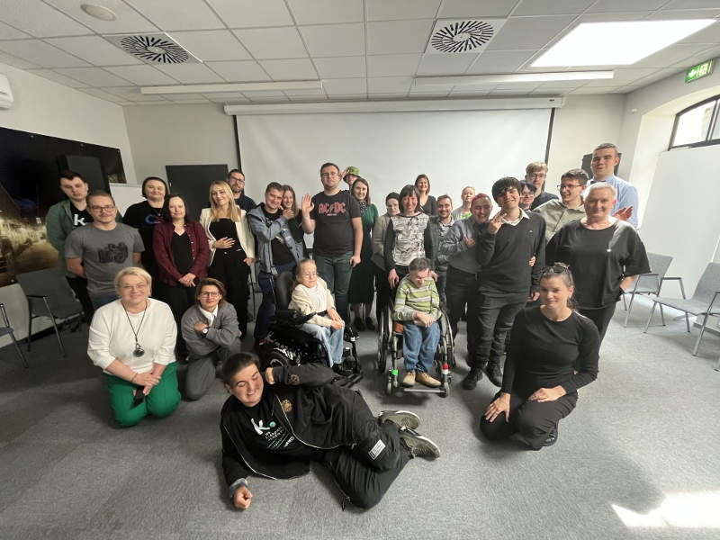 Duża grupa ludzi, w tym osoby na wózkach inwalidzkich i jedna osoba leżąca na podłodze, pozuje do zdjęcia grupowego w jasnym pomieszczeniu.
