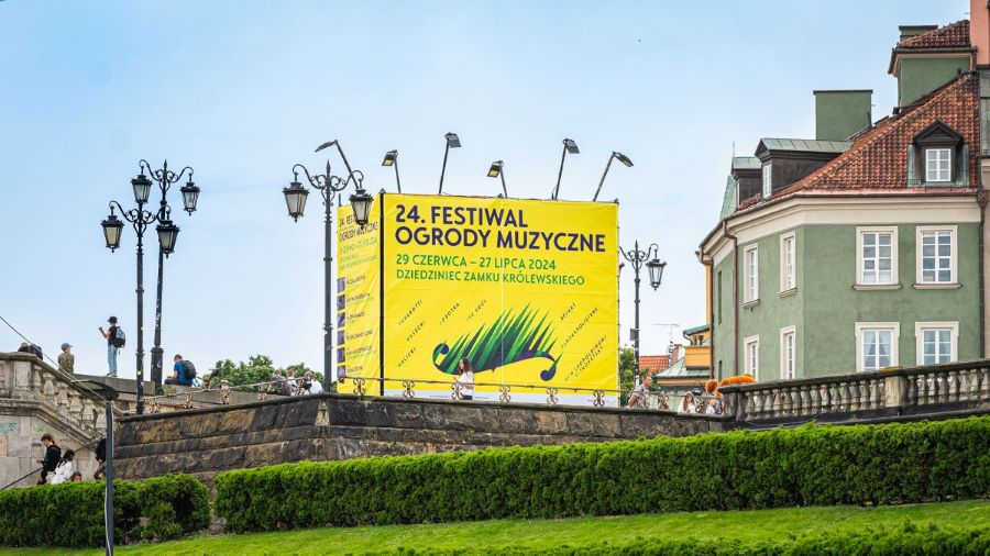 Widok fragmentu placu Zamkowego w Warszawie. Na środku stoi duży billboard z żółtym plakatem reklamującym festiwal.