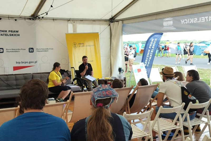 W namiocie osoby, siedzą tyłem do zdjęcia, a przodem do prowadzących. W tle kobieta i mężczyzna prowadzą warszaty. Znajdują się na tle żółtego baneru z logiem PFRON. 