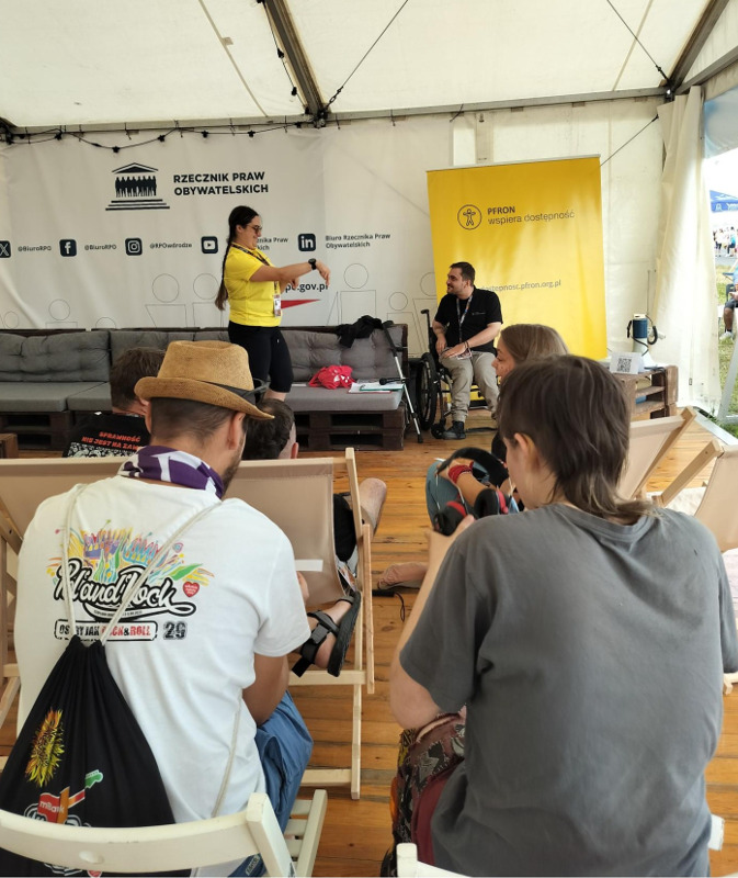 W namiocie osoby, siedzą tyłem do zdjęcia, a przodem do prowadzących. W tle kobieta i mężczyzna prowadzą warszaty. Znajdują się na tle żółtego baneru z logiem PFRON. 