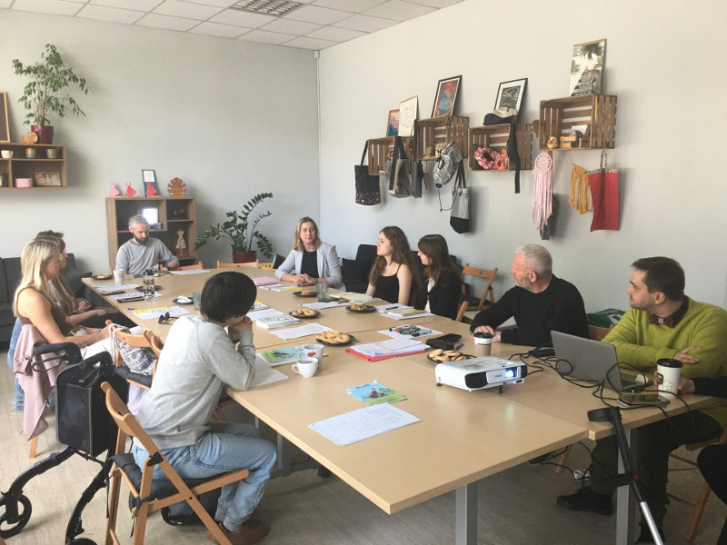 Jasny pokój z długim stołem, przy którym siedzi osiem osób w różnym wieku, w trakcie spotkania lub warsztatów, z laptopami, projektorem i przekąskami na stole.