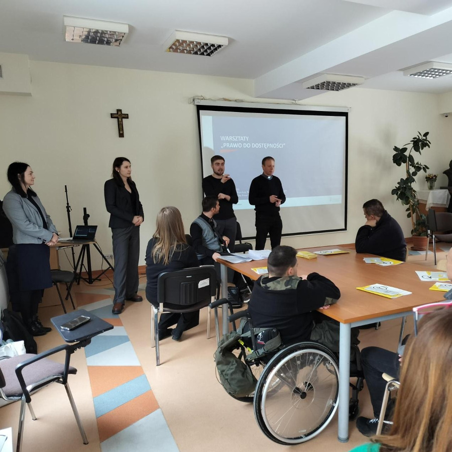 grupa osób, które siedzą przy stole podczas drugiej części spotkania poświęconej pracy warsztatowej. Uczestnicy wspólnie analizują dokumenty i robią notatki, a w tle widoczny jest ekran projekcyjny z napisem „II część praca warsztatowe”.