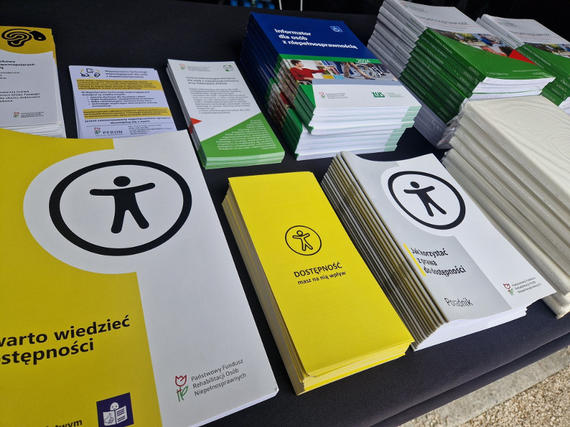 Na czarnym stole leżą stosy informacyjnych broszur i ulotek w różnych kolorach, dotyczących dostępności i praw osób z niepełnosprawnościami, z widocznym logo PFRON