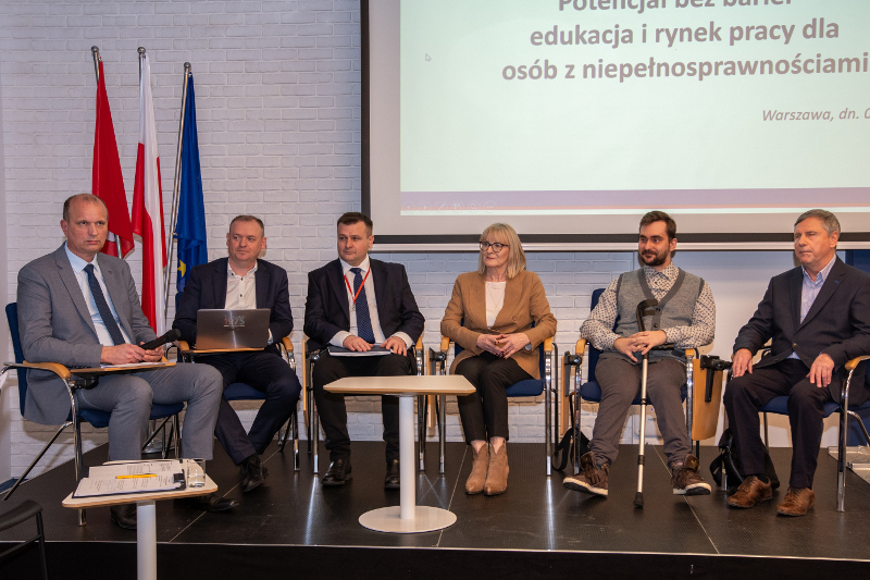 Sześciu panelistów na scenie podczas konferencji (kobieta i pięciu mężczyzn), siedzą na krzesłach, ubrani formalnie. Jeden z panelistów z laską wspierającą. W tle z lewej strony trzy flagi - czerwoną, polską oraz unijną – oraz fragment ekranu z prezentacją zatytułowaną „Potencjał bez barier – edukacja i rynek pracy dla osób z niepełnosprawnościami”. Paneliści patrzą w stronę publiczności.