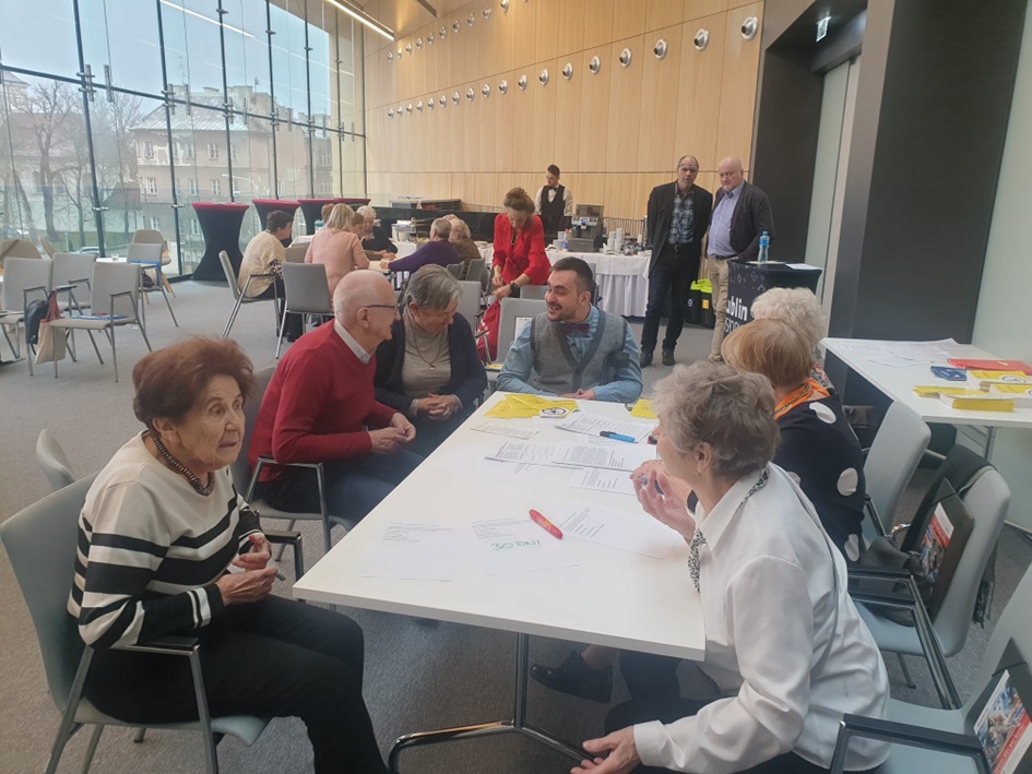 Sala konferencyjna z dużymi oknami. Przy stole siedzi grupa osób pracujących z dokumentami. W tle widoczne są inne osoby i baner „Lublin Gospodarczy”. Zdjęcie przedstawia warsztaty lub konsultacje.