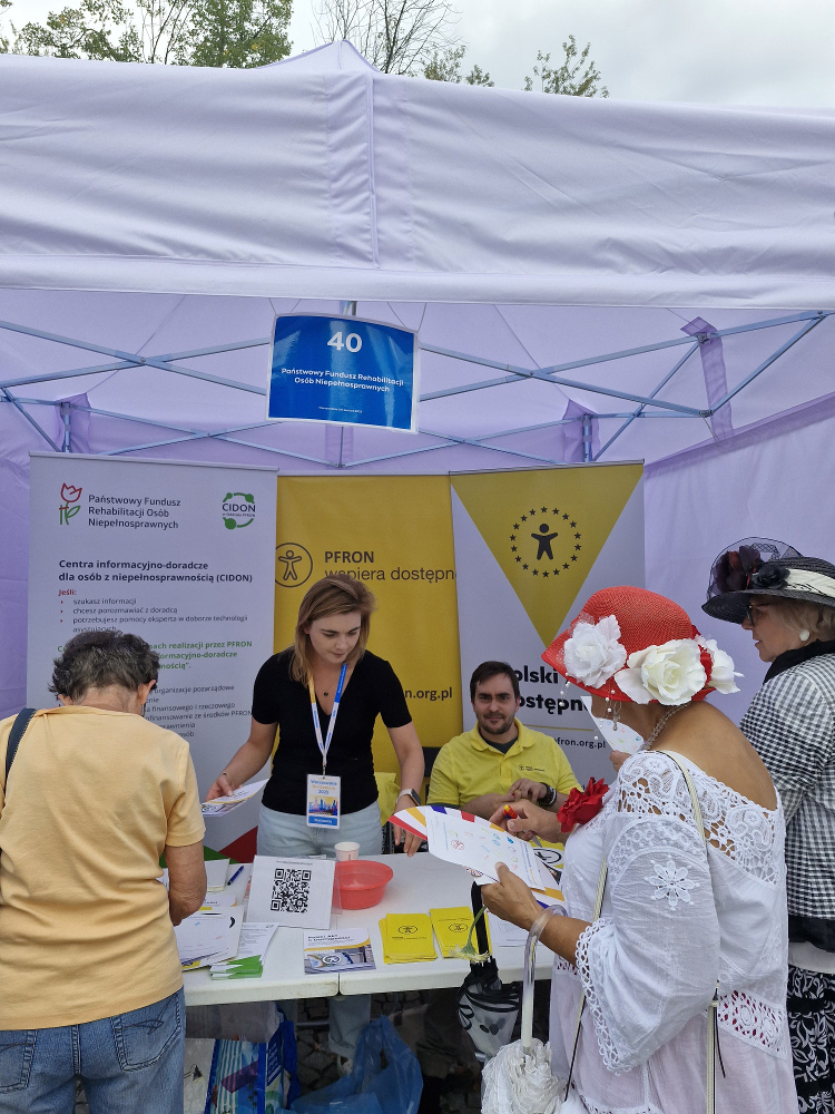Kilka osób stoi przy stoisku i przegląda publikacje PFRON. Stoisko obsługuje kobieta w czarnej koszulce, na szyi ma identyfikator PRON oraz mężczyzna, który ubrany w żółty t-shirt. W tle stoją roll upy z logotypami PFRON.