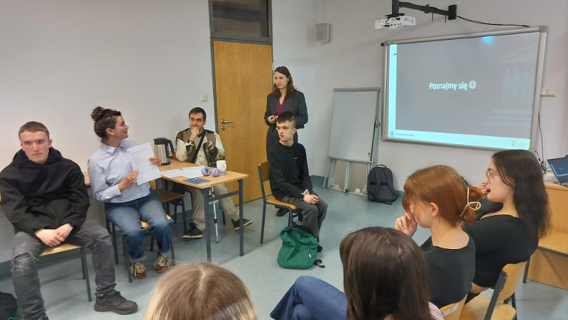 Zdjęcie przedstawia uczestników warsztatów zgromadzonych w sali dydaktycznej podczas wspólnej dyskusji nad dokumentami. Grupa osób, wśród których są studenci ze szczególnymi potrzebami, aktywnie analizuje materiały szkoleniowe w otoczeniu sprzyjającym integracji i praktycznej nauce.