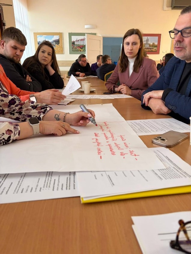 rupa osób przy stole podczas warsztatów, z dokumentami i dużym arkuszem papieru, na którym ktoś pisze czerwonym markerem. W tle obrazy na ścianach oraz uczestnicy skupieni na dyskusji - atmosferę współpracy i wymiany pomysłów.