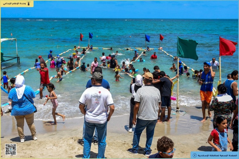 Na brzegu morza stoi kilka osób odwróconych tyłem. W głębi kilkanaście osób niewidomych kąpie się na oznakowanym specjalnymi znacznikami kąpielisku. Słoneczna, letnia pogoda.