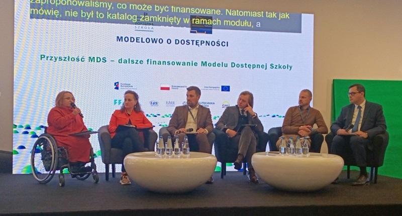 Uczestnicy (6 osób) siedzą przy stolikach z wodą mineralną podczas rozmowy. Małgorzata Radziszewska z PFRON prezentuje temat. W tle plansza informacyjna z tytułem konferencji i panelu: „Modelowo o dostępności. Przyszłość MDS – dalsze finansowanie Modelu Dostępnej Szkoły”.