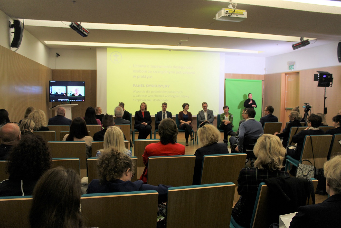 W dolnym rogu widownia (tyłem) patrząca w kierunku panelistów siedzących na tle rzutnika.
